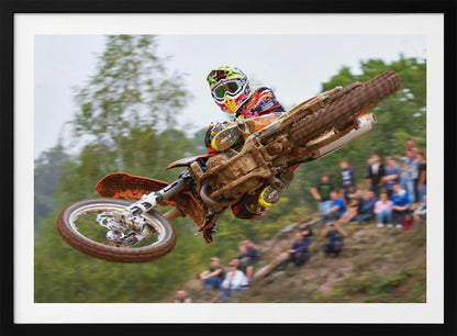A dynamic action shot of a motocross racer in mid-air, leaning into a turn with their muddy orange dirt bike. The rider wears a colorful helmet and racing gear, with a blurred background of green trees and spectators watching from a dirt hill. The photo is enclosed in a silver frame. Decor