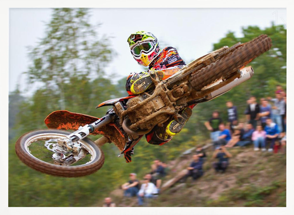 A dynamic action shot of a motocross racer in mid-air, leaning into a turn with their muddy orange dirt bike. The rider wears a colorful helmet and racing gear, with a blurred background of green trees and spectators watching from a dirt hill. The photo is enclosed in a silver frame. Decor