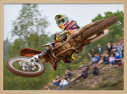 A dynamic action shot of a motocross racer in mid-air, leaning into a turn with their muddy orange dirt bike. The rider wears a colorful helmet and racing gear, with a blurred background of green trees and spectators watching from a dirt hill. The photo is enclosed in a silver frame. Decor