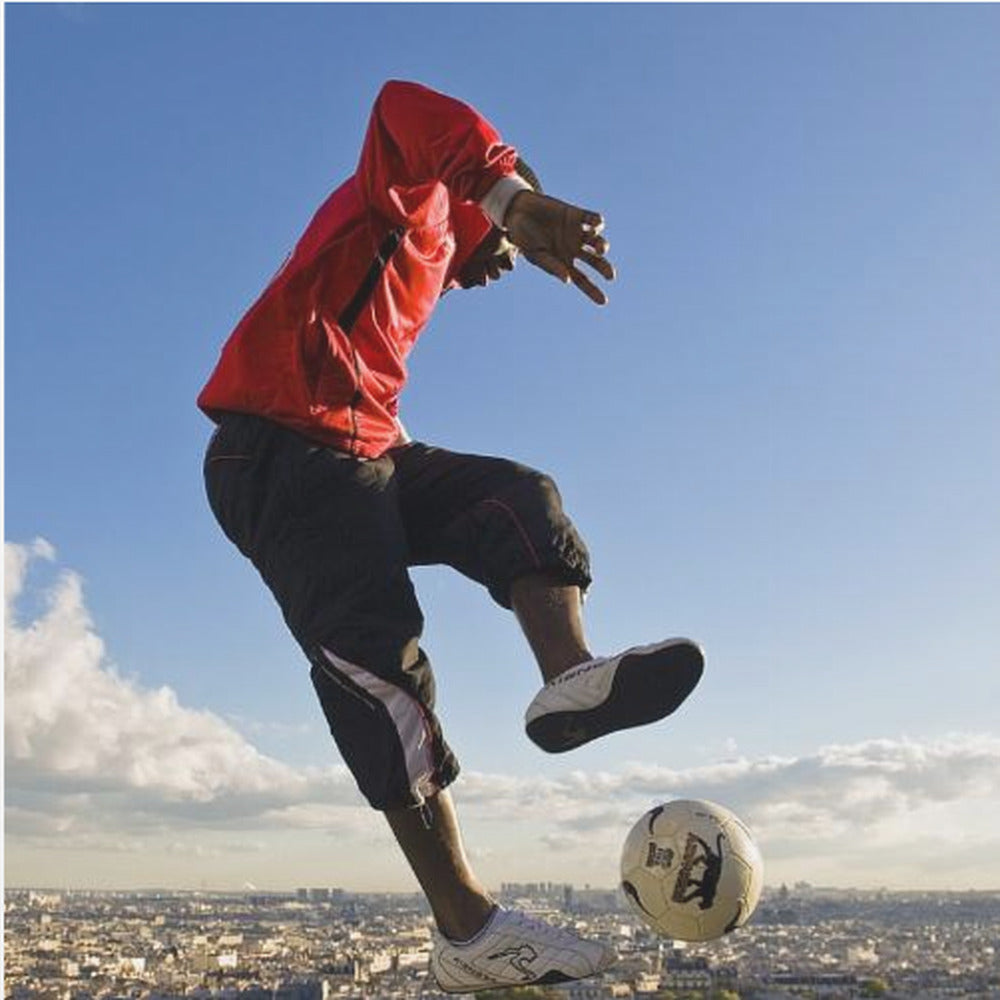 A dynamic low-angle shot of a person in a red jacket and black pants skillfully juggling a soccer ball, suspended in mid-air high above a sprawling city skyline under a clear blue sky, all enclosed within a light wood frame. Artwork