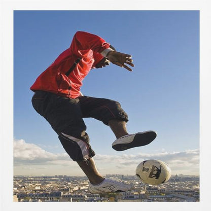 A dynamic low-angle shot of a person in a red jacket and black pants skillfully juggling a soccer ball, suspended in mid-air high above a sprawling city skyline under a clear blue sky, all enclosed within a light wood frame. Artwork