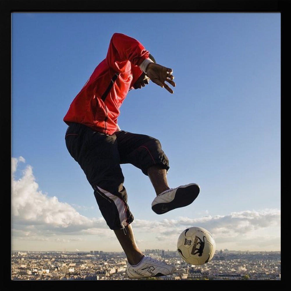 A dynamic low-angle shot of a person in a red jacket and black pants skillfully juggling a soccer ball, suspended in mid-air high above a sprawling city skyline under a clear blue sky, all enclosed within a light wood frame. Artwork