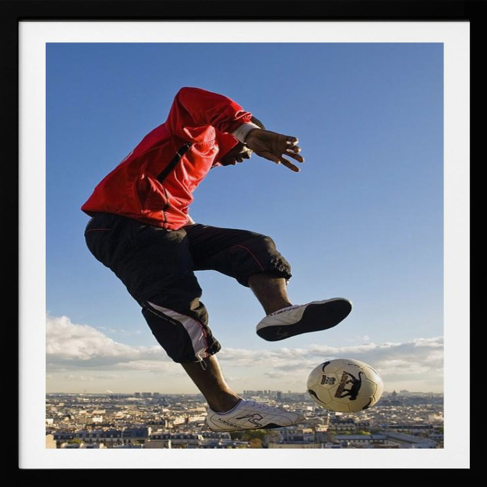 A dynamic low-angle shot of a person in a red jacket and black pants skillfully juggling a soccer ball, suspended in mid-air high above a sprawling city skyline under a clear blue sky, all enclosed within a light wood frame. Artwork
