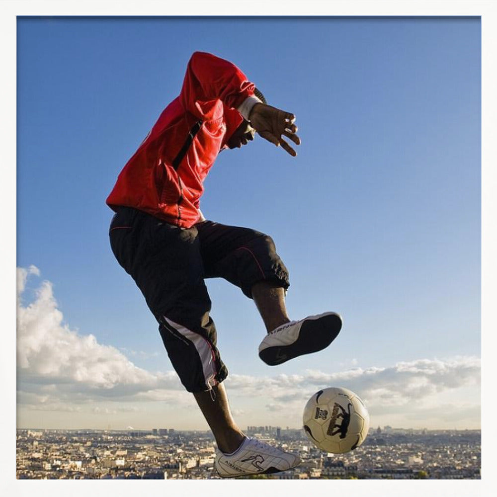 A dynamic low-angle shot of a person in a red jacket and black pants skillfully juggling a soccer ball, suspended in mid-air high above a sprawling city skyline under a clear blue sky, all enclosed within a light wood frame. Artwork