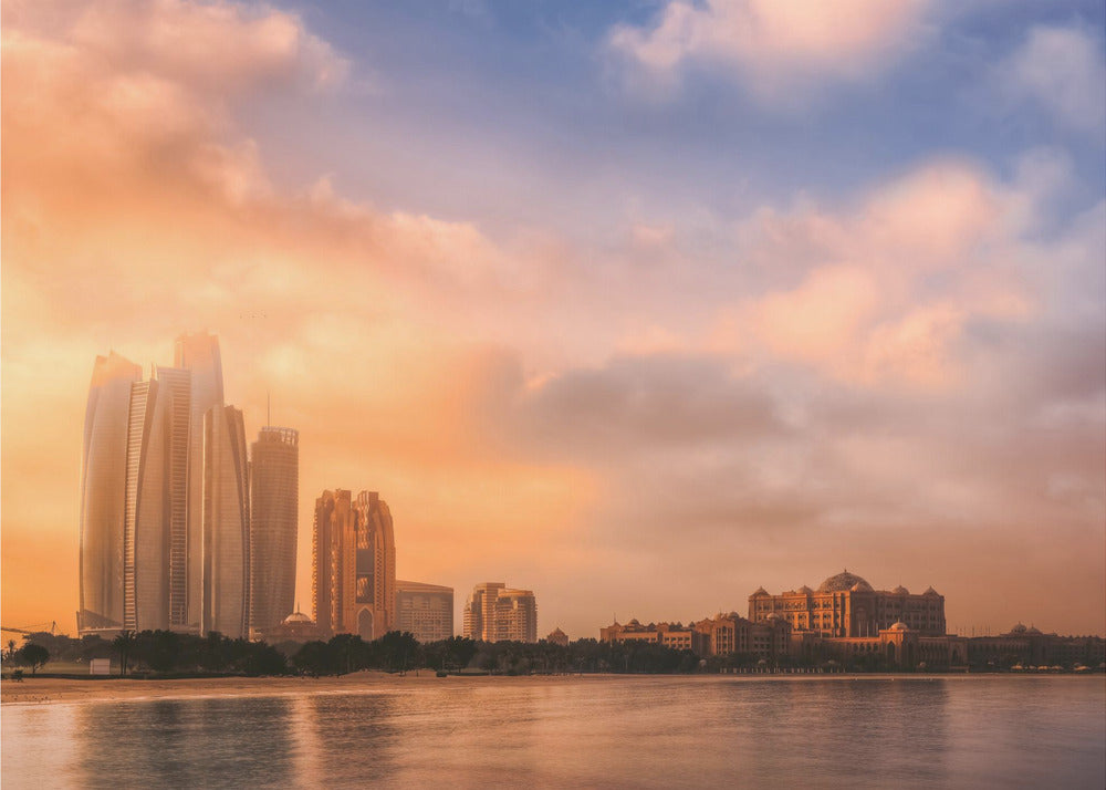 A framed photograph of the Abu Dhabi city skyline at sunset, featuring modern skyscrapers and the Emirates Palace hotel glowing in warm, orange light. The buildings are reflected in the calm water of the foreground, under a sky with soft orange and blue clouds. Poster