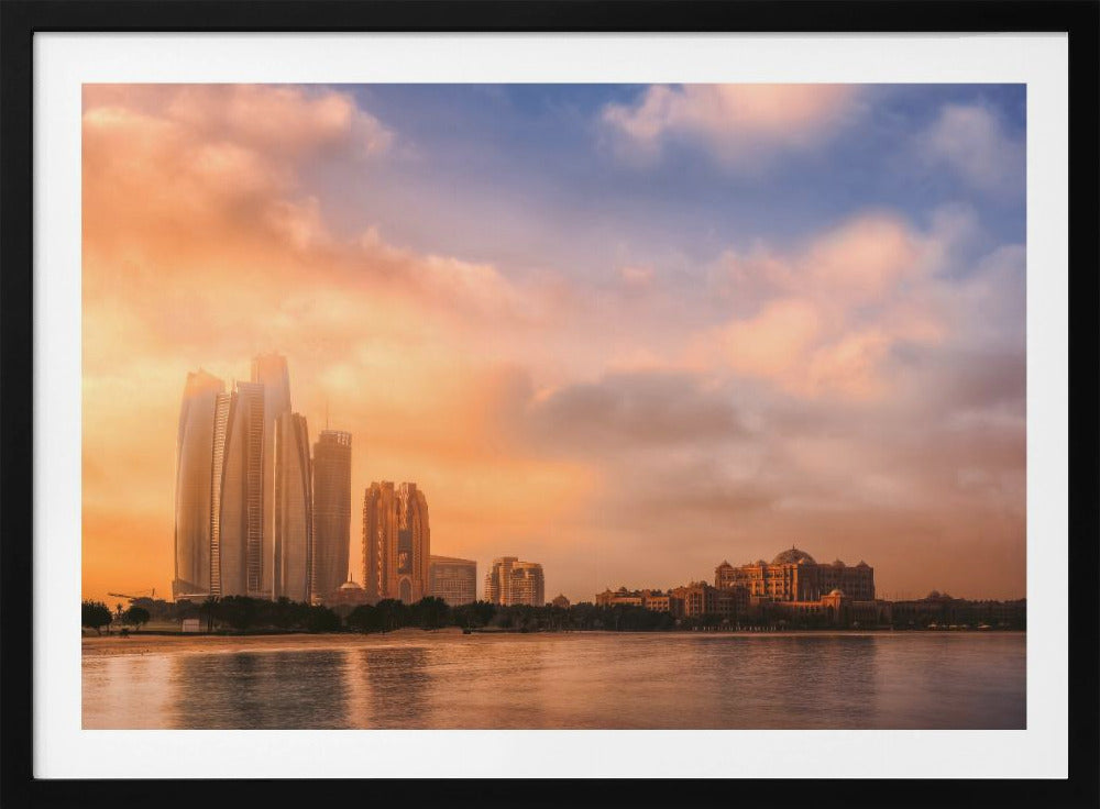 A framed photograph of the Abu Dhabi city skyline at sunset, featuring modern skyscrapers and the Emirates Palace hotel glowing in warm, orange light. The buildings are reflected in the calm water of the foreground, under a sky with soft orange and blue clouds. Poster