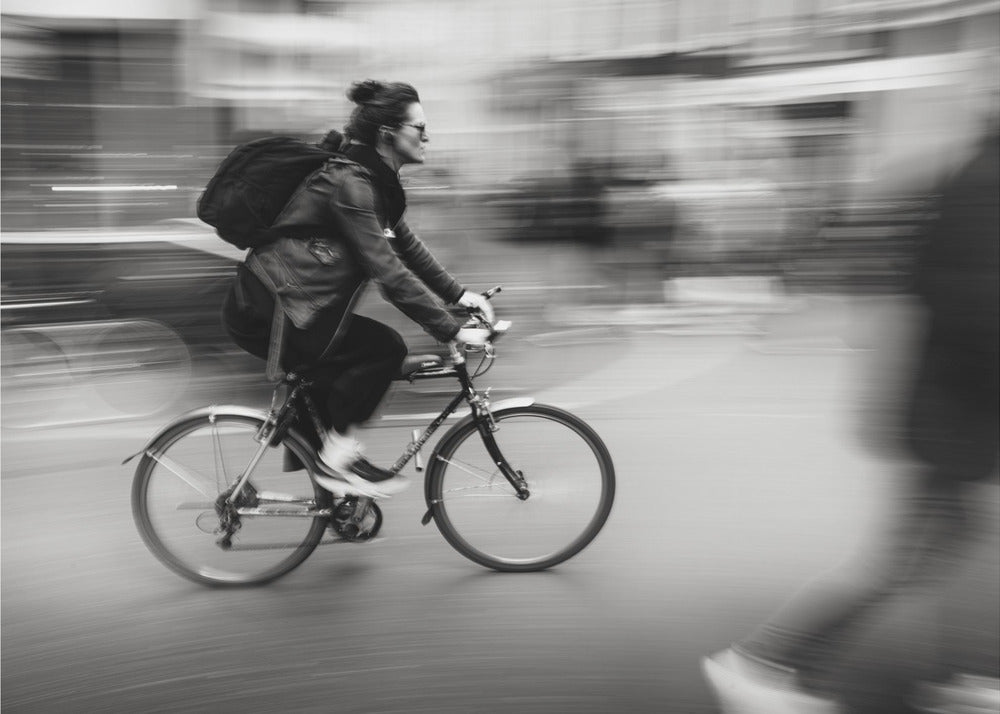 A dynamic black and white photograph capturing a person in profile as they cycle swiftly through a city. The background is a motion blur of urban scenery, highlighting the cyclist's speed. The individual wears a leather jacket, sunglasses, a backpack, and has their hair tied up. The artwork is enclosed in a silver frame. Wall Art