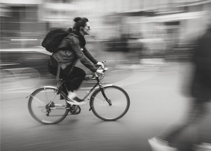 A dynamic black and white photograph capturing a person in profile as they cycle swiftly through a city. The background is a motion blur of urban scenery, highlighting the cyclist's speed. The individual wears a leather jacket, sunglasses, a backpack, and has their hair tied up. The artwork is enclosed in a silver frame. Wall Art