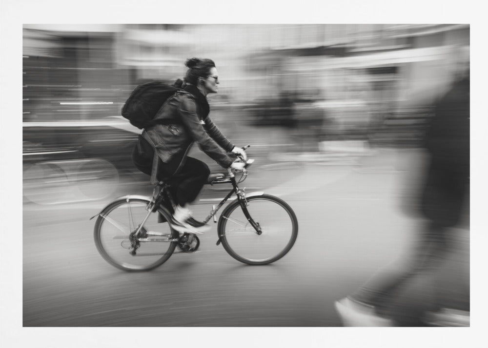 A dynamic black and white photograph capturing a person in profile as they cycle swiftly through a city. The background is a motion blur of urban scenery, highlighting the cyclist's speed. The individual wears a leather jacket, sunglasses, a backpack, and has their hair tied up. The artwork is enclosed in a silver frame. Wall Art