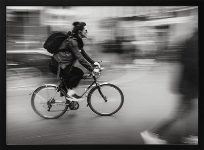 A dynamic black and white photograph capturing a person in profile as they cycle swiftly through a city. The background is a motion blur of urban scenery, highlighting the cyclist's speed. The individual wears a leather jacket, sunglasses, a backpack, and has their hair tied up. The artwork is enclosed in a silver frame. Wall Art
