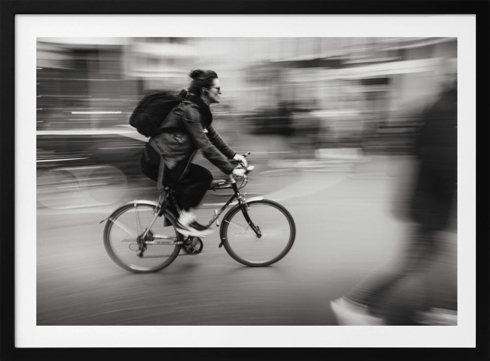A dynamic black and white photograph capturing a person in profile as they cycle swiftly through a city. The background is a motion blur of urban scenery, highlighting the cyclist's speed. The individual wears a leather jacket, sunglasses, a backpack, and has their hair tied up. The artwork is enclosed in a silver frame. Wall Art