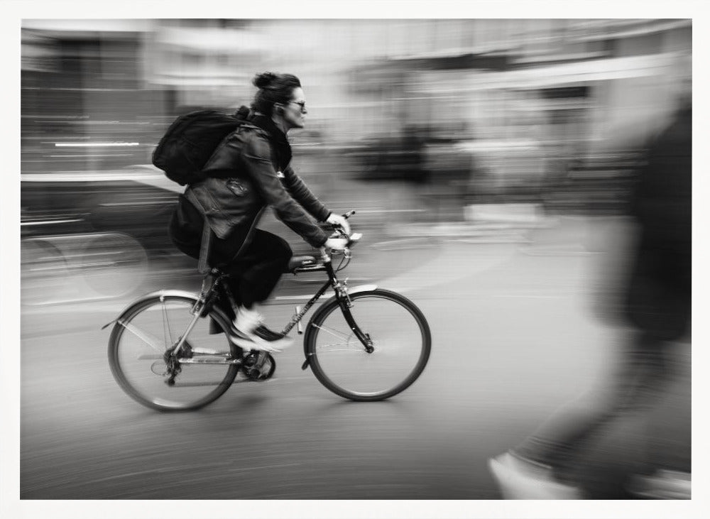 A dynamic black and white photograph capturing a person in profile as they cycle swiftly through a city. The background is a motion blur of urban scenery, highlighting the cyclist's speed. The individual wears a leather jacket, sunglasses, a backpack, and has their hair tied up. The artwork is enclosed in a silver frame. Wall Art