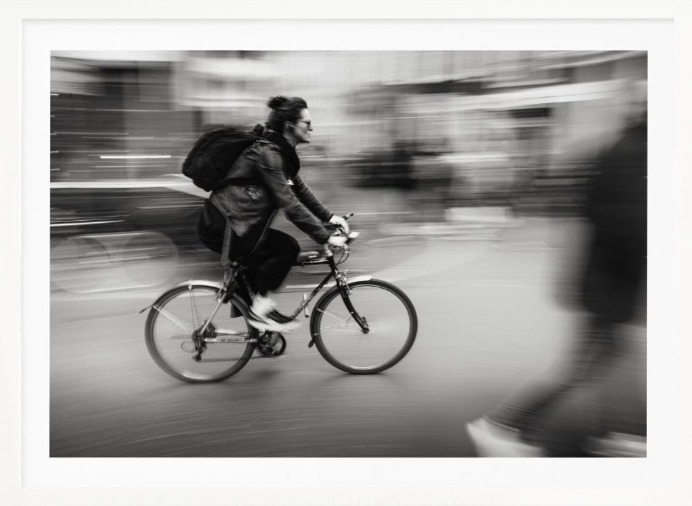 A dynamic black and white photograph capturing a person in profile as they cycle swiftly through a city. The background is a motion blur of urban scenery, highlighting the cyclist's speed. The individual wears a leather jacket, sunglasses, a backpack, and has their hair tied up. The artwork is enclosed in a silver frame. Wall Art