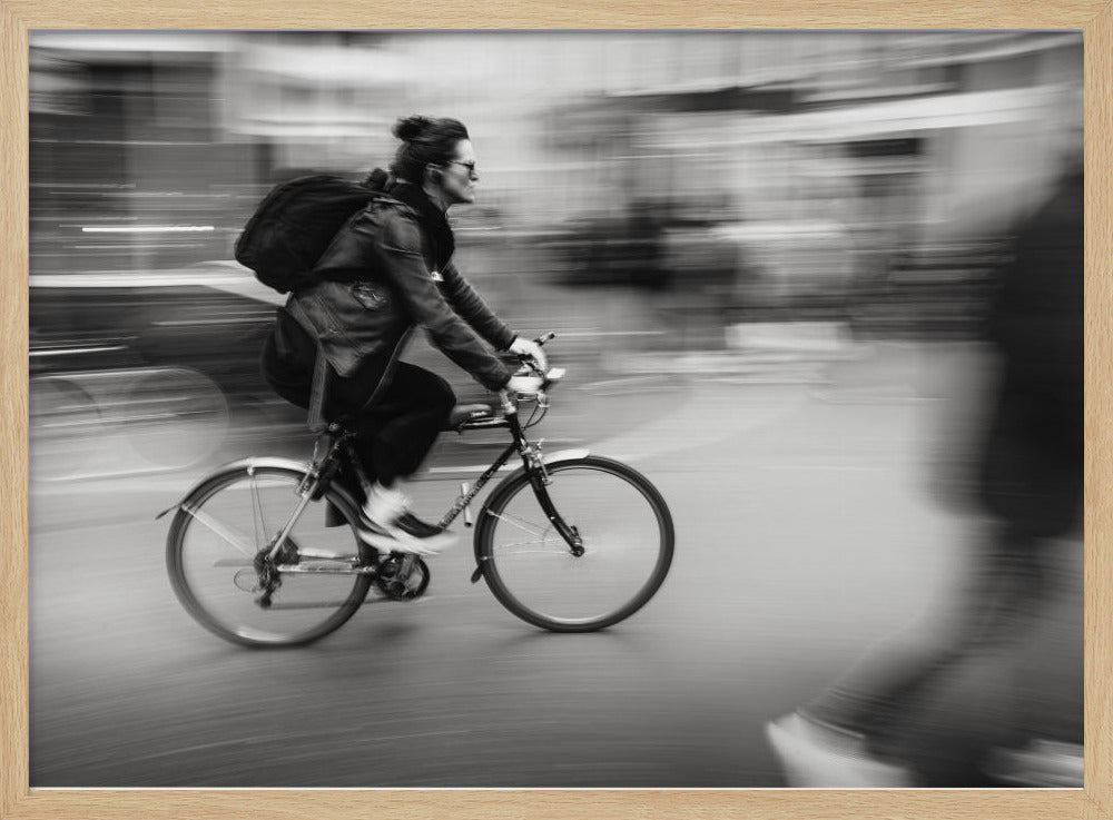 A dynamic black and white photograph capturing a person in profile as they cycle swiftly through a city. The background is a motion blur of urban scenery, highlighting the cyclist's speed. The individual wears a leather jacket, sunglasses, a backpack, and has their hair tied up. The artwork is enclosed in a silver frame. Wall Art