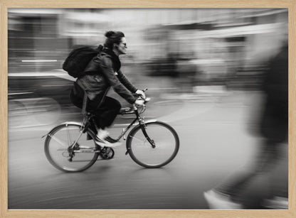 A dynamic black and white photograph capturing a person in profile as they cycle swiftly through a city. The background is a motion blur of urban scenery, highlighting the cyclist's speed. The individual wears a leather jacket, sunglasses, a backpack, and has their hair tied up. The artwork is enclosed in a silver frame. Wall Art