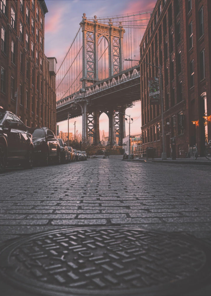 A low-angle photograph taken from a cobblestone street in Dumbo, Brooklyn, looking up towards the Manhattan Bridge at sunset. Red brick buildings frame the view on both sides of the street, with parked cars lining the left. A manhole cover is prominent in the foreground, and the sky is filled with warm pink and orange tones. Print
