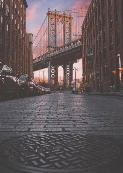 A low-angle photograph taken from a cobblestone street in Dumbo, Brooklyn, looking up towards the Manhattan Bridge at sunset. Red brick buildings frame the view on both sides of the street, with parked cars lining the left. A manhole cover is prominent in the foreground, and the sky is filled with warm pink and orange tones. Print