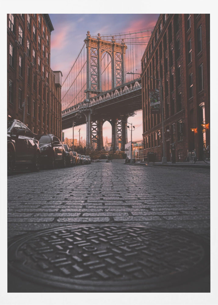 A low-angle photograph taken from a cobblestone street in Dumbo, Brooklyn, looking up towards the Manhattan Bridge at sunset. Red brick buildings frame the view on both sides of the street, with parked cars lining the left. A manhole cover is prominent in the foreground, and the sky is filled with warm pink and orange tones. Print
