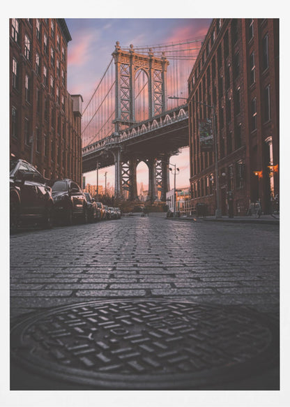 A low-angle photograph taken from a cobblestone street in Dumbo, Brooklyn, looking up towards the Manhattan Bridge at sunset. Red brick buildings frame the view on both sides of the street, with parked cars lining the left. A manhole cover is prominent in the foreground, and the sky is filled with warm pink and orange tones. Print