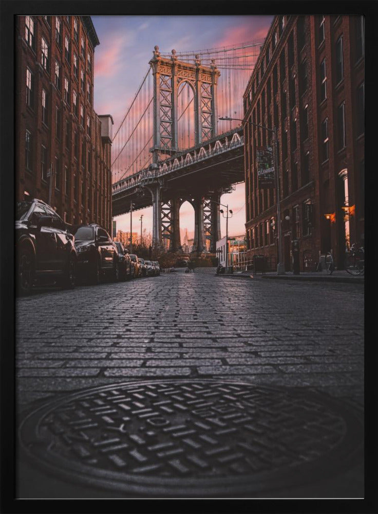 A low-angle photograph taken from a cobblestone street in Dumbo, Brooklyn, looking up towards the Manhattan Bridge at sunset. Red brick buildings frame the view on both sides of the street, with parked cars lining the left. A manhole cover is prominent in the foreground, and the sky is filled with warm pink and orange tones. Print