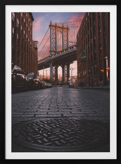 A low-angle photograph taken from a cobblestone street in Dumbo, Brooklyn, looking up towards the Manhattan Bridge at sunset. Red brick buildings frame the view on both sides of the street, with parked cars lining the left. A manhole cover is prominent in the foreground, and the sky is filled with warm pink and orange tones. Print