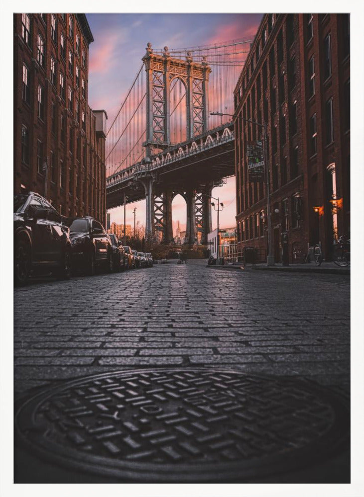 A low-angle photograph taken from a cobblestone street in Dumbo, Brooklyn, looking up towards the Manhattan Bridge at sunset. Red brick buildings frame the view on both sides of the street, with parked cars lining the left. A manhole cover is prominent in the foreground, and the sky is filled with warm pink and orange tones. Print