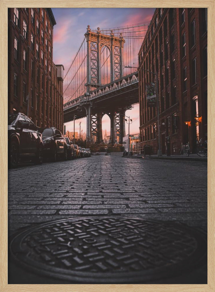 A low-angle photograph taken from a cobblestone street in Dumbo, Brooklyn, looking up towards the Manhattan Bridge at sunset. Red brick buildings frame the view on both sides of the street, with parked cars lining the left. A manhole cover is prominent in the foreground, and the sky is filled with warm pink and orange tones. Print