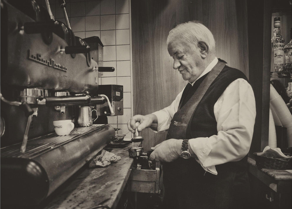 A sepia-toned, framed photograph of an elderly barista with white hair, wearing a vest, as he skillfully prepares an espresso. He is focused on his task, tamping the coffee grounds with a gentle smile, standing next to a large, vintage-style espresso machine in a cozy cafe setting. Print