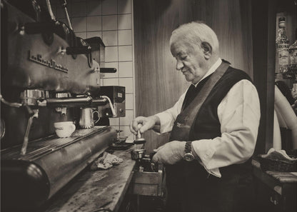 A sepia-toned, framed photograph of an elderly barista with white hair, wearing a vest, as he skillfully prepares an espresso. He is focused on his task, tamping the coffee grounds with a gentle smile, standing next to a large, vintage-style espresso machine in a cozy cafe setting. Print