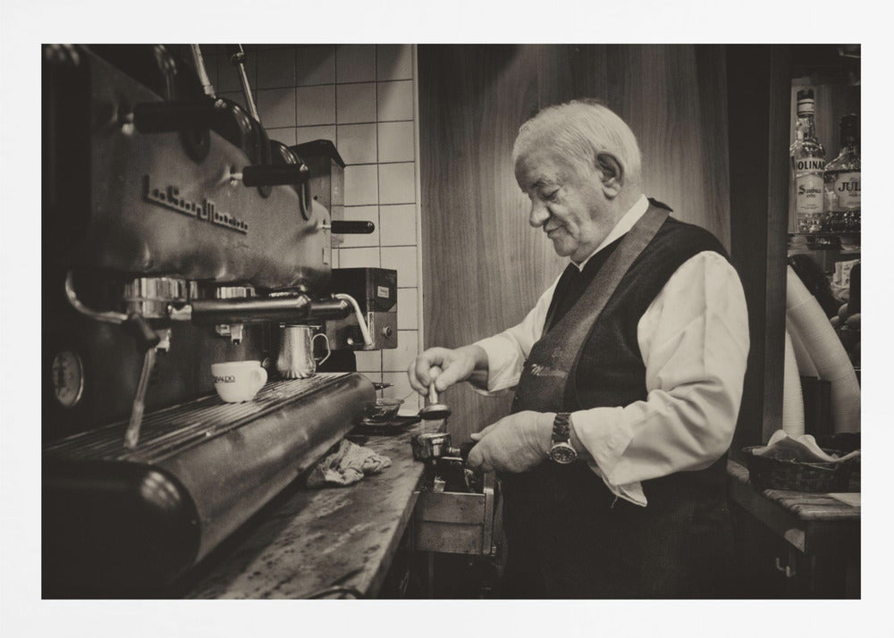 A sepia-toned, framed photograph of an elderly barista with white hair, wearing a vest, as he skillfully prepares an espresso. He is focused on his task, tamping the coffee grounds with a gentle smile, standing next to a large, vintage-style espresso machine in a cozy cafe setting. Print