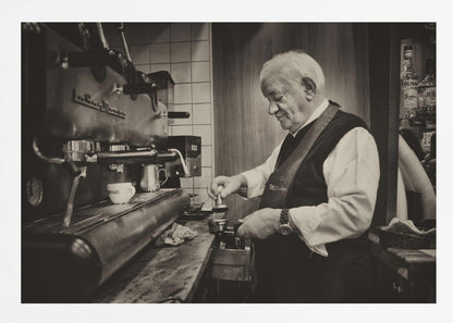 A sepia-toned, framed photograph of an elderly barista with white hair, wearing a vest, as he skillfully prepares an espresso. He is focused on his task, tamping the coffee grounds with a gentle smile, standing next to a large, vintage-style espresso machine in a cozy cafe setting. Print