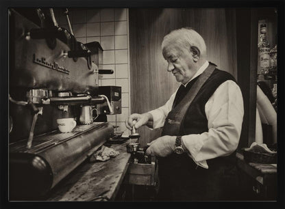 A sepia-toned, framed photograph of an elderly barista with white hair, wearing a vest, as he skillfully prepares an espresso. He is focused on his task, tamping the coffee grounds with a gentle smile, standing next to a large, vintage-style espresso machine in a cozy cafe setting. Print