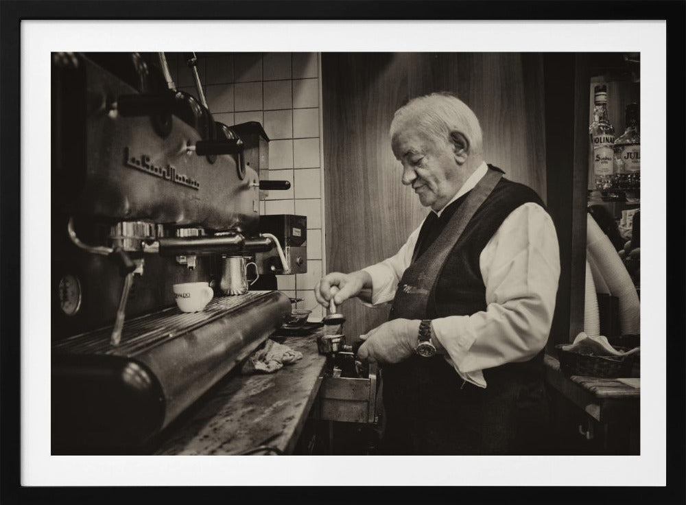 A sepia-toned, framed photograph of an elderly barista with white hair, wearing a vest, as he skillfully prepares an espresso. He is focused on his task, tamping the coffee grounds with a gentle smile, standing next to a large, vintage-style espresso machine in a cozy cafe setting. Print