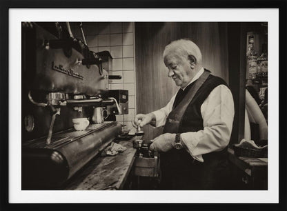 A sepia-toned, framed photograph of an elderly barista with white hair, wearing a vest, as he skillfully prepares an espresso. He is focused on his task, tamping the coffee grounds with a gentle smile, standing next to a large, vintage-style espresso machine in a cozy cafe setting. Print