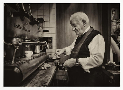 A sepia-toned, framed photograph of an elderly barista with white hair, wearing a vest, as he skillfully prepares an espresso. He is focused on his task, tamping the coffee grounds with a gentle smile, standing next to a large, vintage-style espresso machine in a cozy cafe setting. Print