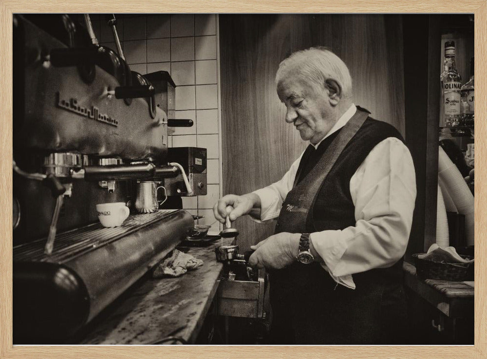 A sepia-toned, framed photograph of an elderly barista with white hair, wearing a vest, as he skillfully prepares an espresso. He is focused on his task, tamping the coffee grounds with a gentle smile, standing next to a large, vintage-style espresso machine in a cozy cafe setting. Print