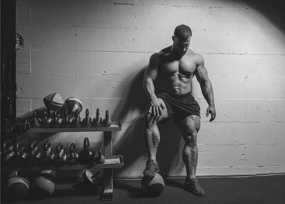 A dramatic black and white photograph of a shirtless, muscular bodybuilder posing in a gym. He leans against a cinder block wall with one foot propped on a medicine ball, his chiseled physique and defined muscles highlighted by intense lighting. Racks of kettlebells are visible in the background. Wall Art