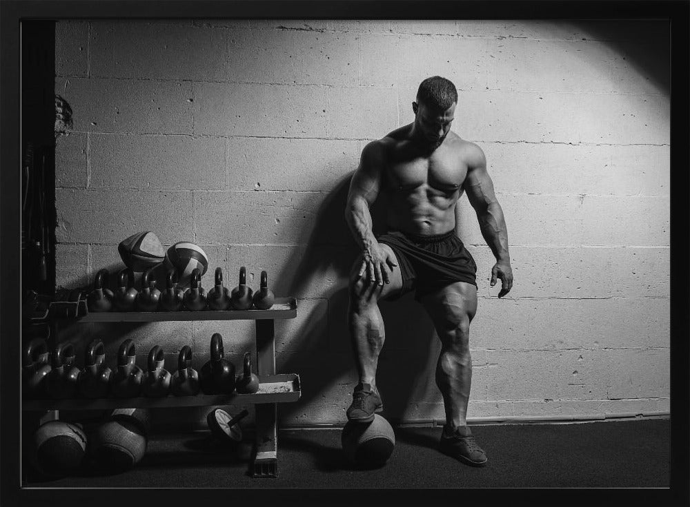 A dramatic black and white photograph of a shirtless, muscular bodybuilder posing in a gym. He leans against a cinder block wall with one foot propped on a medicine ball, his chiseled physique and defined muscles highlighted by intense lighting. Racks of kettlebells are visible in the background. Wall Art