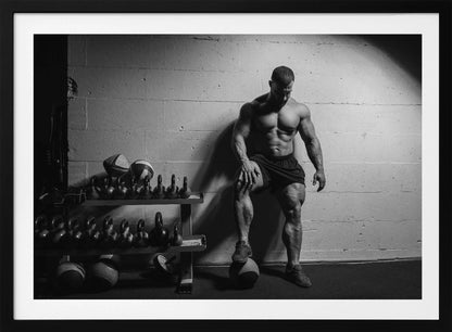 A dramatic black and white photograph of a shirtless, muscular bodybuilder posing in a gym. He leans against a cinder block wall with one foot propped on a medicine ball, his chiseled physique and defined muscles highlighted by intense lighting. Racks of kettlebells are visible in the background. Wall Art