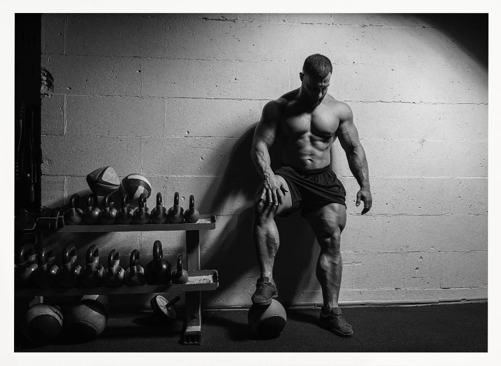 A dramatic black and white photograph of a shirtless, muscular bodybuilder posing in a gym. He leans against a cinder block wall with one foot propped on a medicine ball, his chiseled physique and defined muscles highlighted by intense lighting. Racks of kettlebells are visible in the background. Wall Art