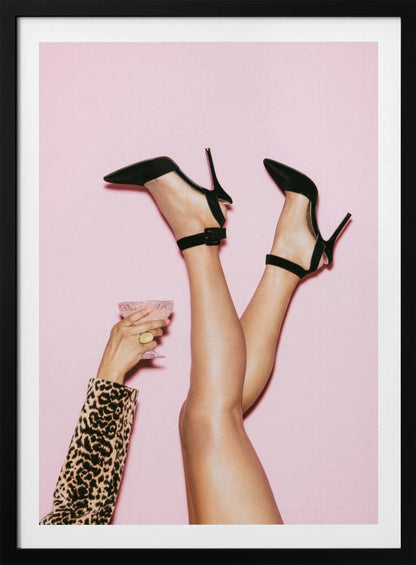 A woman's legs are kicked up in the air against a solid pink background. She is wearing black, pointed-toe, high-heeled shoes with ankle straps. A hand in a leopard print sleeve holds a pink, textured cocktail glass to the side. Artwork