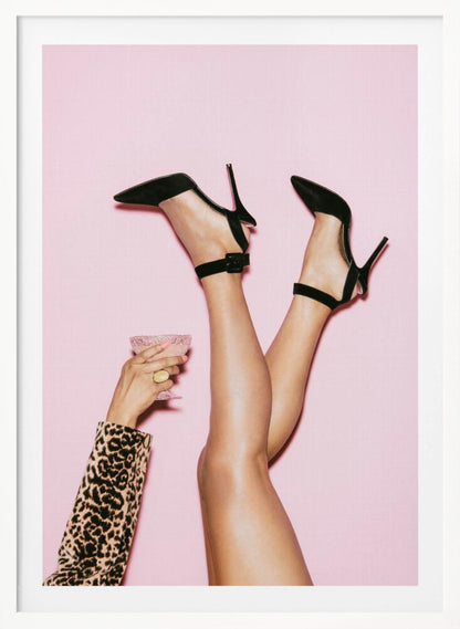 A woman's legs are kicked up in the air against a solid pink background. She is wearing black, pointed-toe, high-heeled shoes with ankle straps. A hand in a leopard print sleeve holds a pink, textured cocktail glass to the side. Artwork