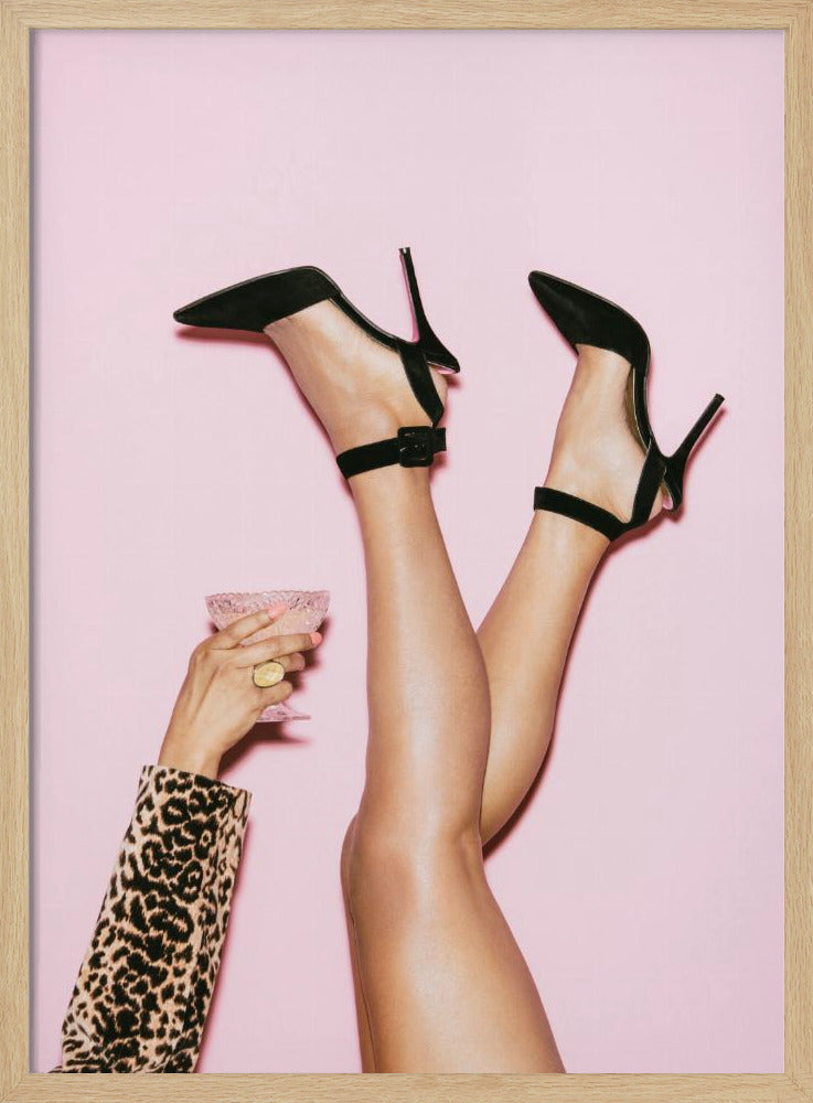 A woman's legs are kicked up in the air against a solid pink background. She is wearing black, pointed-toe, high-heeled shoes with ankle straps. A hand in a leopard print sleeve holds a pink, textured cocktail glass to the side. Artwork
