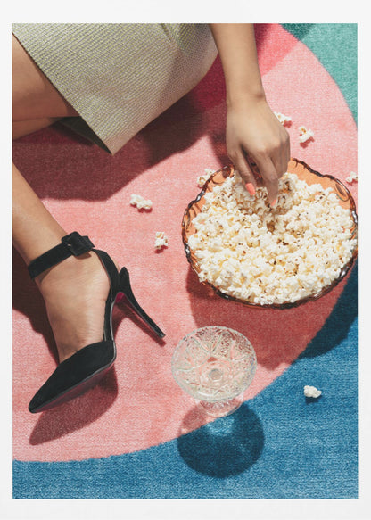 A high-angle photograph of a woman sitting on a vibrant pink and blue rug. She is wearing a chic black stiletto high heel and reaching into a glass bowl filled with popcorn. A crystal glass and scattered popcorn kernels are also on the rug. Artwork