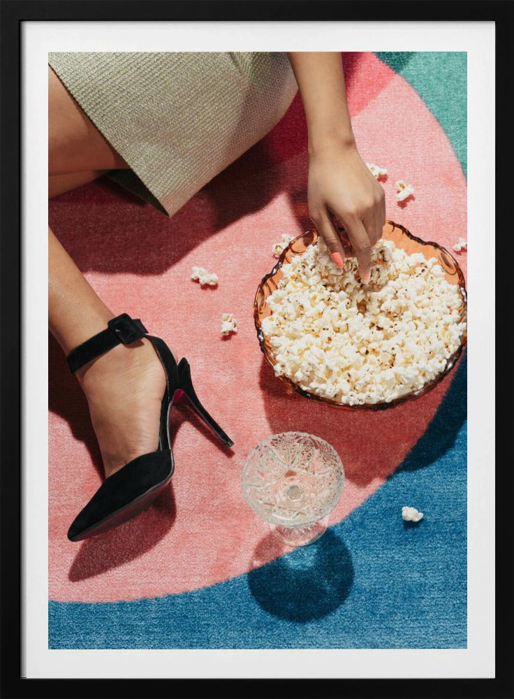 A high-angle photograph of a woman sitting on a vibrant pink and blue rug. She is wearing a chic black stiletto high heel and reaching into a glass bowl filled with popcorn. A crystal glass and scattered popcorn kernels are also on the rug. Artwork