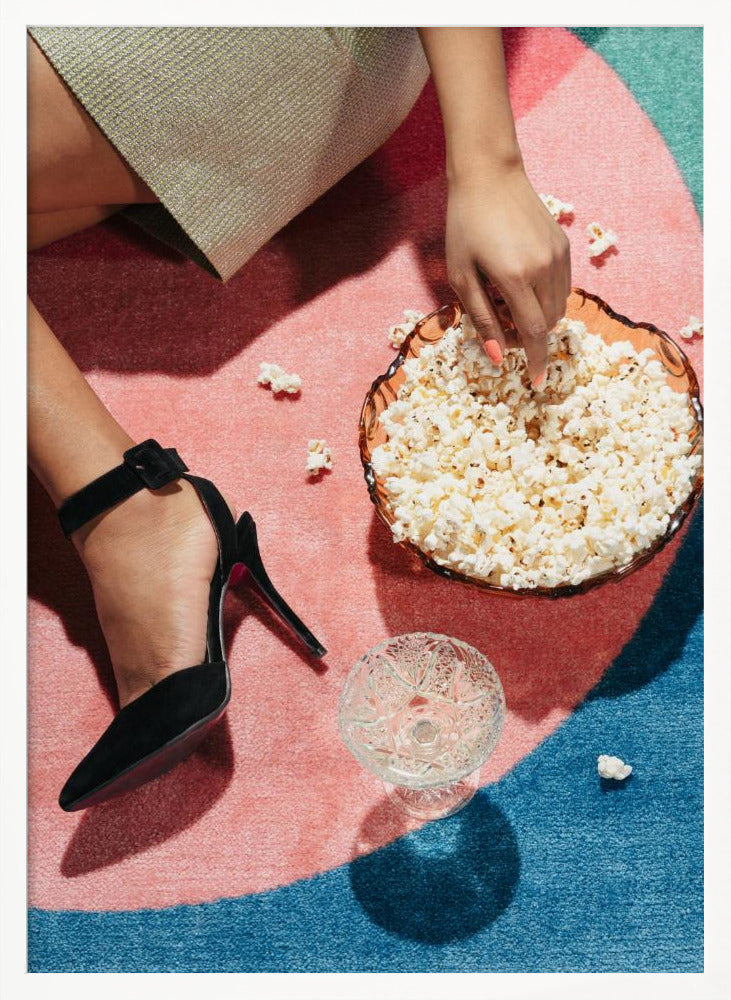 A high-angle photograph of a woman sitting on a vibrant pink and blue rug. She is wearing a chic black stiletto high heel and reaching into a glass bowl filled with popcorn. A crystal glass and scattered popcorn kernels are also on the rug. Artwork