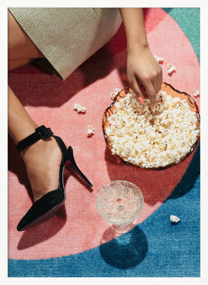 A high-angle photograph of a woman sitting on a vibrant pink and blue rug. She is wearing a chic black stiletto high heel and reaching into a glass bowl filled with popcorn. A crystal glass and scattered popcorn kernels are also on the rug. Artwork