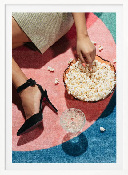 A high-angle photograph of a woman sitting on a vibrant pink and blue rug. She is wearing a chic black stiletto high heel and reaching into a glass bowl filled with popcorn. A crystal glass and scattered popcorn kernels are also on the rug. Artwork