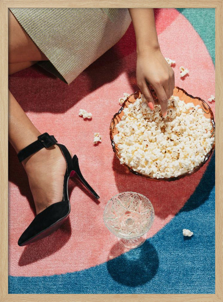 A high-angle photograph of a woman sitting on a vibrant pink and blue rug. She is wearing a chic black stiletto high heel and reaching into a glass bowl filled with popcorn. A crystal glass and scattered popcorn kernels are also on the rug. Artwork