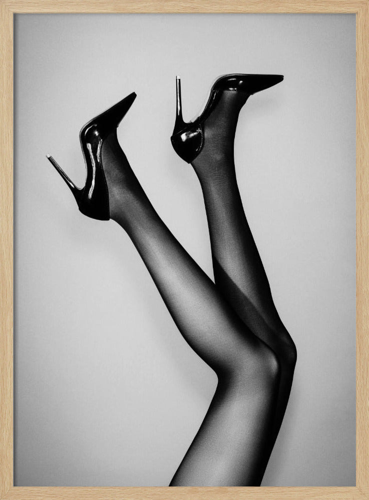 A black and white photograph of a woman's legs in the air, wearing sheer black stockings and shiny black stiletto high heels against a plain grey background. Poster